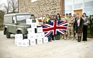 Wendy Clarkson Fizzco’s company director standing with her team and some Army personnel holding a British flag in front of Fizzco’s s original headquarters. To the left is a series 2 Land Rover, in front of the vehicle, is a pile of white boxes filled with Christmas decorations.